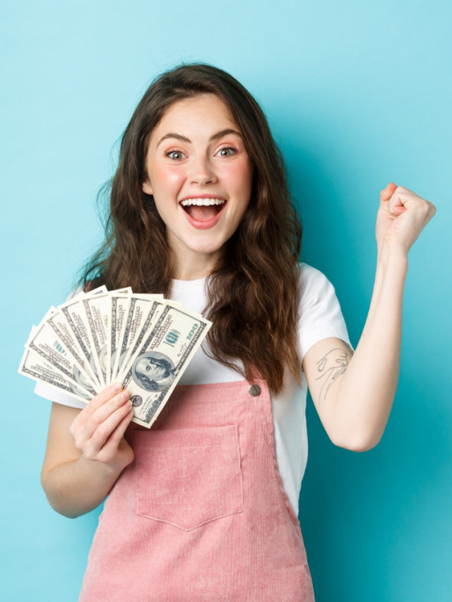 cropped excited smiling girl fist pump hold money prize winning cash receive income from something standing happy against blue background